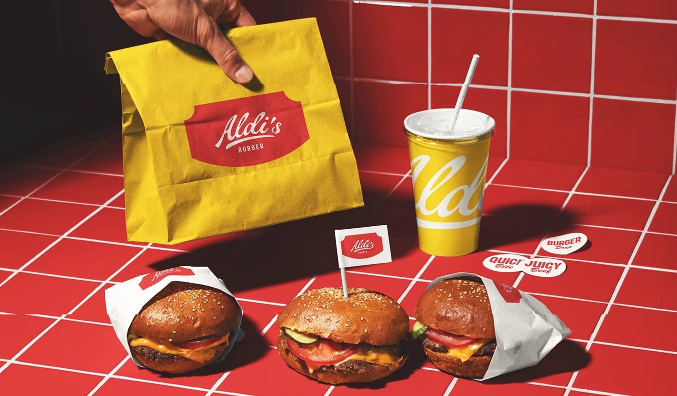 Aldi's Burger Visual Identity — 노란 종이봉투와 컵, 빨간 타일 위 버거들. 봉투에는 배지 하나