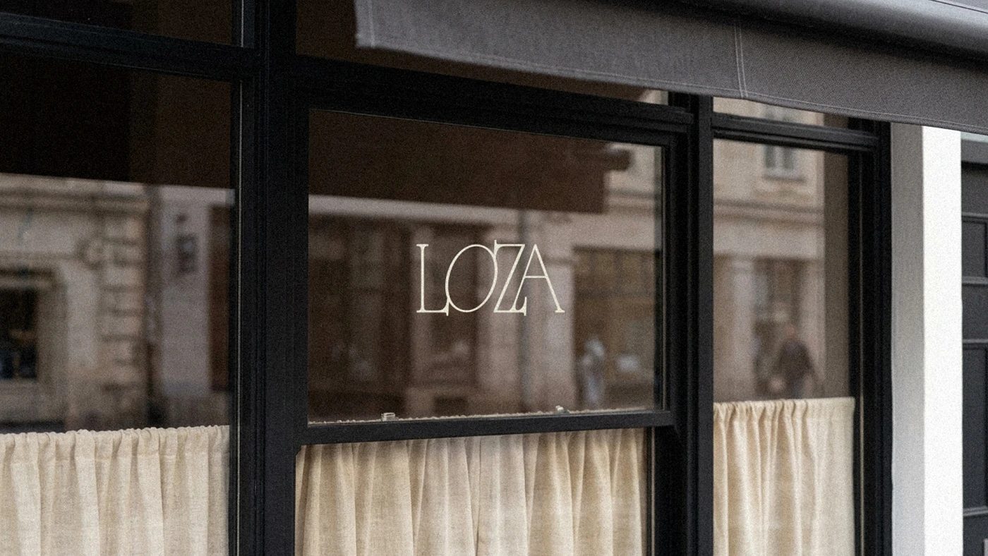 loza / restaurant — 창문. 유리에 가는 세리프 로고 LOZA, 안쪽은 반만 가린 리넨 커튼